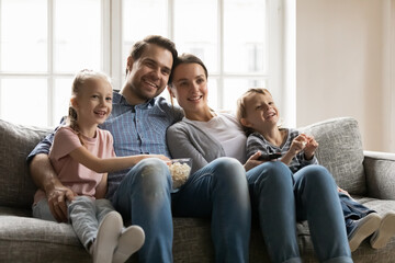 Happy bonding family of four resting on comfortable sofa, watching movie together indoors. Smiling young parents cuddling little children siblings, enjoying funny TV series with popcorn at home.