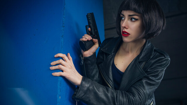 Beautiful brunette sexy spy agent (killer or police) woman in leather jacket and jeans with a gun in her hand running after someone, to catch him on parking