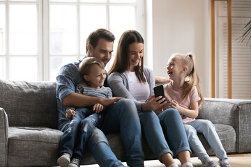 Obraz premium Smiling young woman showing funny video on smartphone to laughing little children and handsome father, relaxing together on comfortable sofa in living room, family weekend hobby pastime concept.