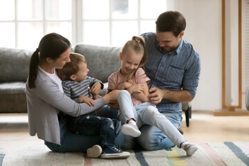 Full length playful young parents tickling laughing little daughter and son, enjoying spending leisure time together at home. Happy couple having fun with small kids, playing on floor in living room.