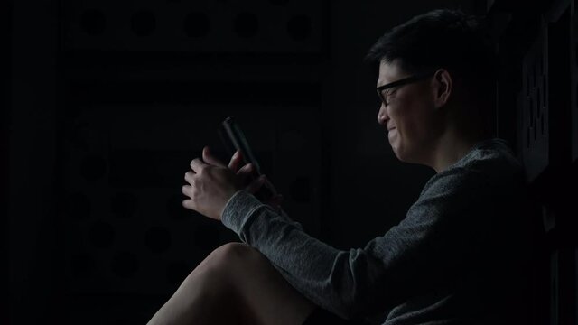 sad young asian man sitting on the floor checking mobile phone with bad news message in the dark at home he lost job in quarantine while COVID-19 or  Coronavirus Pandemic .