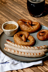 Bavarian traditional grilled pork sausages on ceramic plate served with german sweet mustard, mug of dark beer and pretzels bread on white and blue napkin over wooden background.