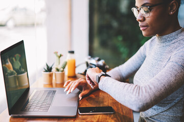 Prosperous afro american businesswoman in trendy eyewear checking time while waiting for payment confirmation making transaction online,female owner of company looking at watch waiting for meeting