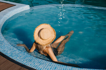 Young woman sitting in a pool, wearing a straw hat on a sunny day.