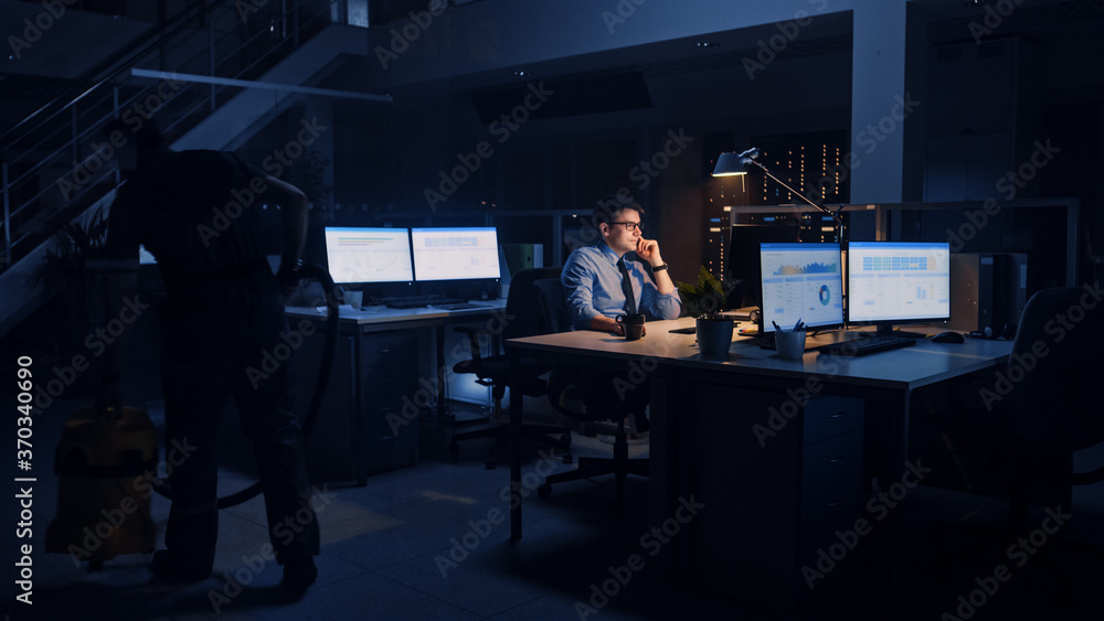 Poster Working Late in Office: Businessman Uses Desktop Computer, Analyzing, Using Documents, Solving Problems, Finishing Important Project. Diligent Ambitious Young Worker. Cleaning Man Vacuums Floor