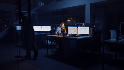 Working Late at Night in Office: Businessman Uses Desktop Computer, Analyzing, Using Documents, Solving Problems, Finishing Important Project. Ambitious Young Worker. Cleaning Man Swipes Floor