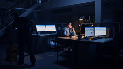 Working Late in Office: Businessman Uses Desktop Computer, Analyzing, Using Documents, Solving Problems, Finishing Important Project. Diligent Ambitious Young Worker. Cleaning Man Vacuums Floor