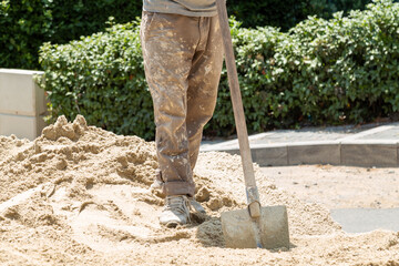 Construction worker in dirty uniform holding in his hand a shovel standing on a pile of sand and is...
