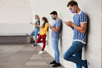 Young people wearing face safety masks using smart mobile phones while keeping social distance on...