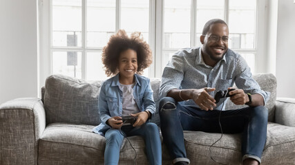 Overjoyed young biracial father sit on sofa have fun play video games with cute little daughter,...