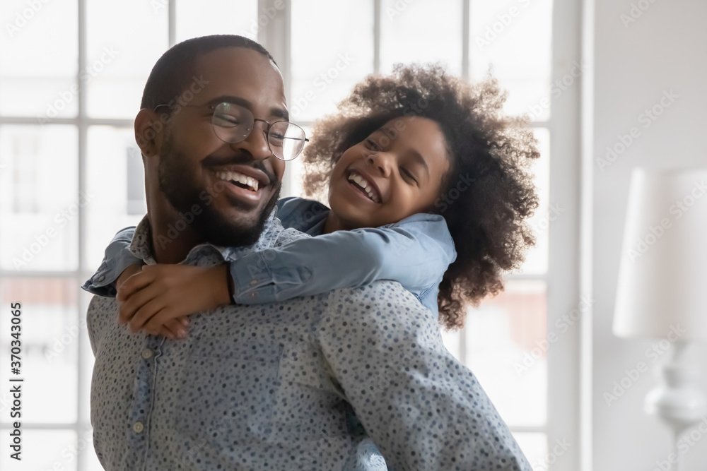 Wall mural excited little ethnic daughter child piggyback smiling young african american father playing at home