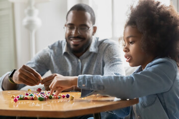 Little biracial girl child and loving young father string colorful wooden beads on thread have fun together at home, loving african American dad and daughter make bracelets, creative activity concept