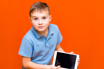 Portrait of european schoolboy with tablet and blue t-shirt on orange studio background.