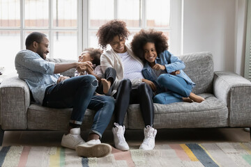 Overjoyed young biracial parents relax on sofa have fun with small ethnic children son and daughter, happy african American family with little kids play rest on couch at home, tickle entertain