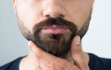 Close up of young bearded man touching his beard while standing against gray background