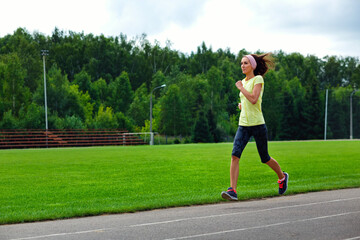 Fototapeta premium A beautiful fitness girl in a yellow sports top and leggings is Jogging around the stadium. She runs fast on a warm summer day. The Sportswoman Is Engaged In Her Usual Sports Practice.