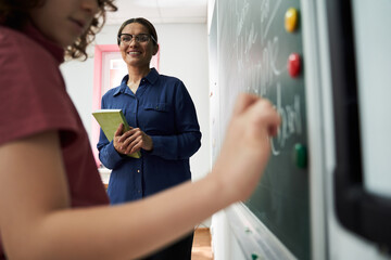 female teacher watching pupil standing at blackboard