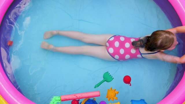 Young girl in pink swimsuit playing in colorful rainbow inflatable swimming pool on backyard top view. Summer domestic active lifestyle swim for kid, fun home water leisure for children,hot weather 4K