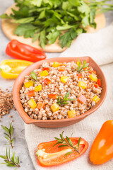Buckwheat porridge with vegetables in clay bowl on a gray concrete background and linen textile. Side view, close up, selective focus.