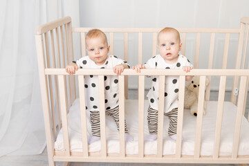 two baby twins brother and sister 8 months sit in their pajamas in the crib and look at the camera,...
