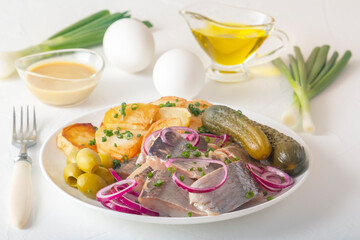 Slices of pickled herring with red onion, fermented cucumber, fried potatoes, olives, mustard sauce, boiled eggs and herbs on a light wooden table. Close-up