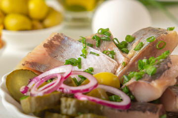 Slices of pickled herring with red onion, fermented cucumber, fried potatoes, olives, boiled eggs and herbs on a light wooden table. Close-up