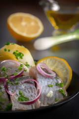 Pickled herring slices with red onion, fermented cucumber, potatoes, olives and lemon in a black plate on a dark wooden table. Close-up