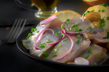 Pickled herring slices with red onion, fermented cucumber, potatoes, olives and lemon in a black plate on a dark wooden table. Close-up