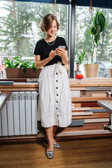 Happy woman standing next to a window, typing in her phone.