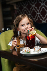 Little girl in overall dress sitting in cafe, drinking juice from a jar