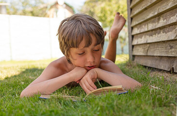 boy reluctantly reads a book lying on the grass in his garden, vacation, back to school, horizontal...