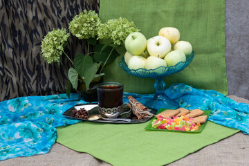 On the table there is a glass of tea in a cup holder, a vase of apples, on a plate are cookies and candied fruits. Nearby is a bouquet of green hydrangea inflorescences.