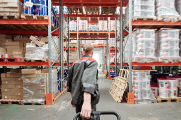 Rear view of young blond male worker of warehouse in uniform pulling loaded cart
