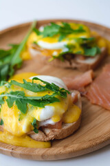 Poached eggs Benedict with Hollandaise sauce, smoked salmon, arugula on toasted bread. Wooden plate, white background.