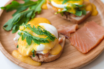 Poached eggs Benedict with Hollandaise sauce, smoked salmon, arugula on toasted bread. Wooden plate, white background.