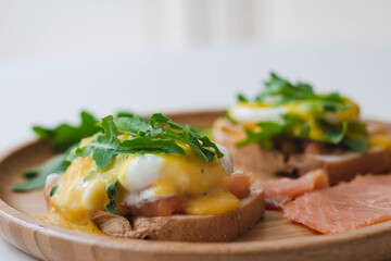 Poached eggs Benedict with Hollandaise sauce, smoked salmon, arugula on toasted bread. Wooden plate, white background.