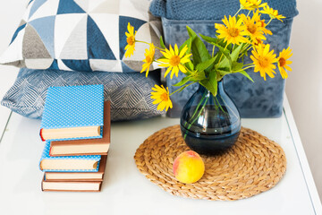 Cozy home interior decor: stack of books, peach, decorative pillows, box with plaid and vase with yellow flowers on a glass table. Distance home education.Quarantine concept of stay home.