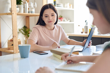 Young mother with laptop looking at her daughter making notes in copybook