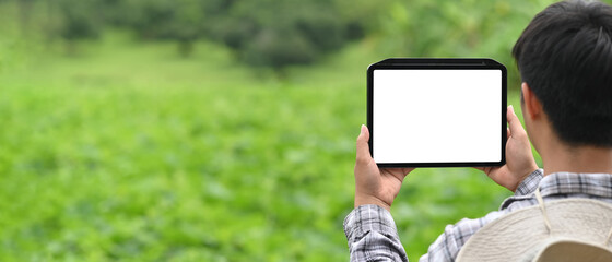 A man is using a white blank screen computer tablet while sitting over the grass field as a background.