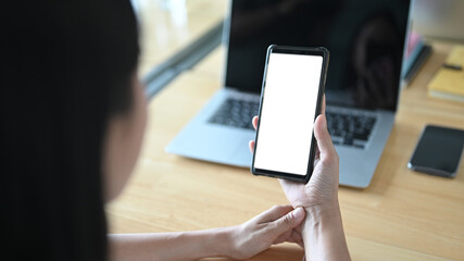 A woman is using a white blank screen mobile phone while sitting in front of a computer laptop at the wooden table.