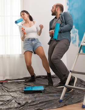 Joyful couple redecorating, dacing and singing with roller brush dipped in blue paint. Apartment redecoration and home construction while renovating and improving. Repair and decorating.