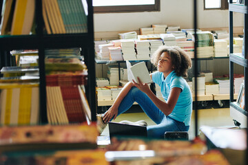 Curly teenage girl sitting on the floor in library and reading interesting book