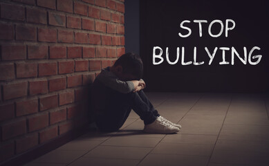 Message STOP BULLYING and sad little boy sitting on floor near brick wall indoors