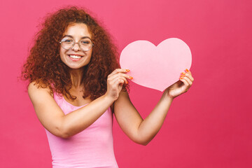 Photo of amazing young curly woman holding big pink paper heart thinking over right answer on date invitation wears casual isolated over pink background.