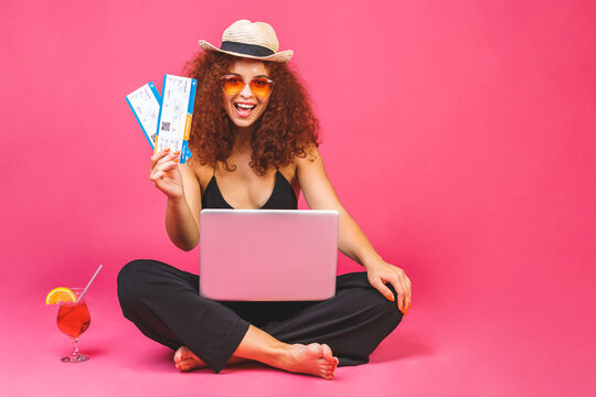 Summer business concept. Portrait of happy woman in casual sitting on floor in lotus pose and holding laptop, cocktail and flight ticket isolated over pink background.