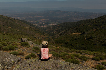 Fototapeta na wymiar Attractive young female seated on the top of a rock witnessing the landscape of the north of Extremadura, Spain while wearing a reflex camera to take photos