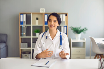 Doctor woman looking at the computer web camera smiling at the patient sitting at the table in the...