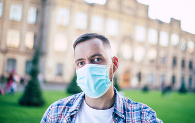 Young handsome confident student man in the medical mask is looking on the camera and standing on university background