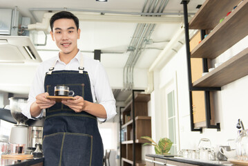 Asian barista man holding a cup of hot black coffee, smile and look at camera in the coffee shop