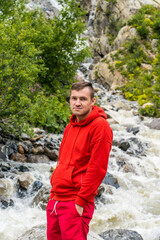 Portrait of young man looking away on background of waterfall. Adult male tourist enjoying beautiful view in mountainous area.